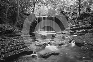 Black and white Epic beautiful Autumn landscape image of Nant Mill waterfall in Wales with glowing sunlight through the woodland