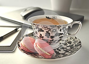 A black and white floral tea cup with pink French macaroons on a table with laptop computer and a mouse - a work from home workst photo