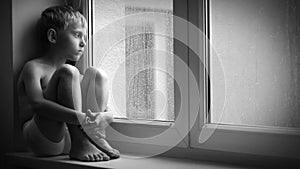 Black and white footage of a sad boy sitting on the windowsill during downpour, unable to get out of the apartment.