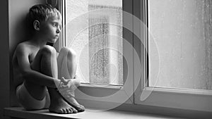 Black and white footage of a sad boy sitting on the windowsill during downpour, unable to get out of the apartment.