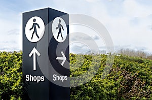 Black and white freestanding directional sign post with arrows and person symbol icon pointing towards shops.