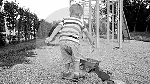 Black and white image of 3 years old toddler boy sitting in the sandbox at palyground and playing with toy truck