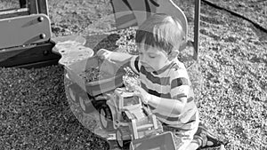 Black and white image of 3 years old toddler boy sitting in the sandbox at palyground and playing with toy truck