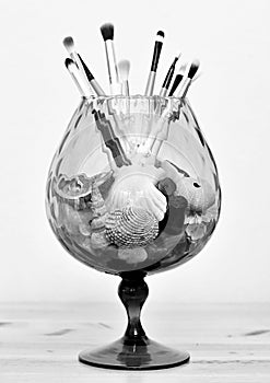 Black and white image of a giant wine glass with makeup brushes standing upright in shells, nail polish and semi-precious stones.