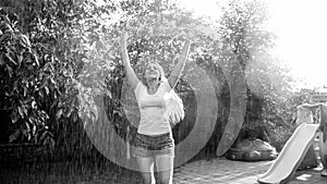 Black and white image of laughing young woman in wet clothes enjoying warm rain at backyard garden. Girl playing and