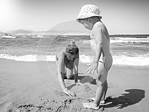 Black and white image of little toddler boy playing with young mother on the sea beach. Family relaxing and having good