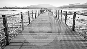 Black and white image of long wooden pier in the ocean. Calm sea waves and amazing sunset over the mountains