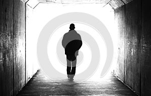 Black and white image of a man hands in pockets walking away in a dark tunnel towards the light.