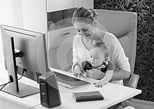 Black and white image of young owman sitting with her baby son behind computer desk at office