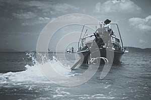 Black and white matt toned. The driver of a longtail motorboat drives a motor with a long drive and a propeller. Sailor in orange
