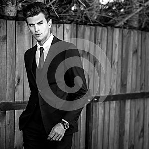 Black-white outdoor portrait of elegant handsome man in classical suit near wooden fence