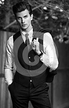 Black-white outdoor portrait of elegant handsome man in classical vest near wooden fence