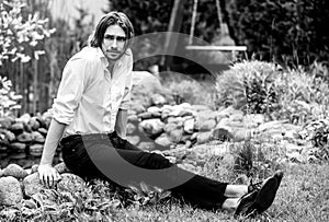 Black-white outdoor portrait of elegant long hair handsome man siting near pond