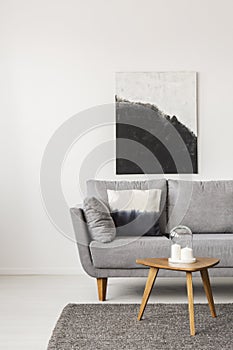 Black and white painting on empty white wall with wooden coffee table with candles and grey sofa with pillow