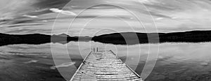Black and white panorama of a calm lake with reflections of mountains and sky and a wooden dock in the foreground