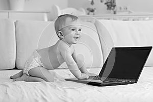 Black and white photo of adorable smiling baby boy sitting on sofa and using laptop