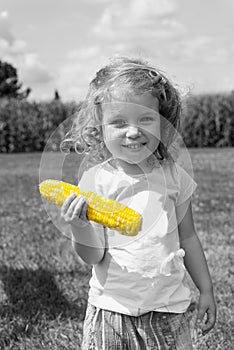 Black and white photo with color corn. Adorable little blonde Caucasian girl is on the field and eating a corn. The stalks of cor