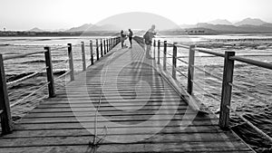 Black and white image of long wooden pier in the ocean. Calm sea waves and amazing sunset over the mountains