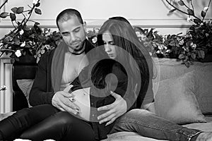 Black and white photo of muslim man and his pregnant wife, he hugging her belly while sitting on the gray cozy