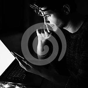 Black and white photo; portrait of thoughtful young man wearing glasses using smartphone and laptop.