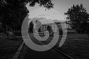 Black and white photo of small town old park plunging in dusk. Outdoor city place for walking with decayed old benches