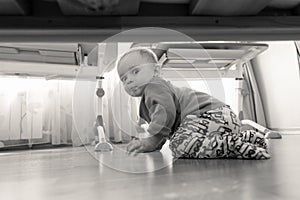 Black and white image of smiling toddler boy sitting on wooden floor at bedroom and looking under the bed