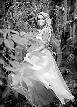 Black and white photo of a stunning girl in a wedding dress, blonde with long curls circling in nature.