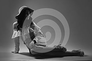 Black and white photo of stylish young woman in hat, shirt and skirt sits on floor over  background.