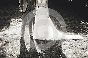 Black and white photo of wedding couple standing on grass in park. Back-light view with shadows pointing to camera