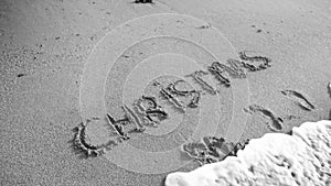 Black and white photo of word Christmas written on wet san on the beach. Sea wave flowing over. Concept of winter