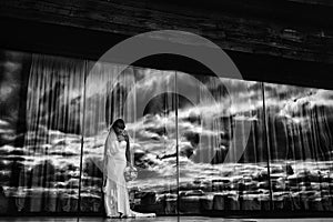 Black and white photo of young bride in white long dress and bouquet of flowers in hand outdoor by the big mirror window