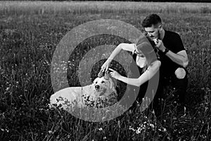 Black and white photo of young pregnant couple who is indulging in walk with their dog, putting children`s shoes on her head.