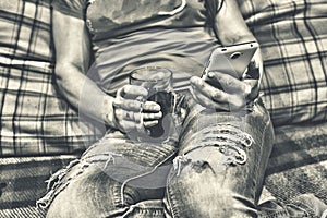 Black and white photo. Young woman in torn jeans is using a white smartphone while sitting on the couch