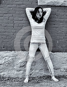 Black and white photograph of female model standing against brick wall with backdrop of old buildings