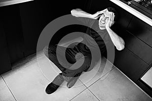 Black and white picture of young man in black clothes sitting separated alone in kitchen and scream. Hold hands on head