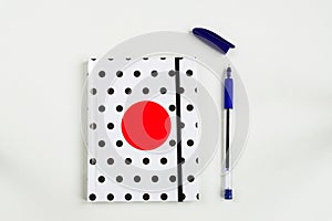 Black and white polka dot note book with red circle  on the cover and blue pen on white table. top view, minimal flat lay