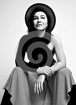 Black and white portrait of adult good looking brunette woman sitting in modern black sleeveless top, maxi skirt and hat