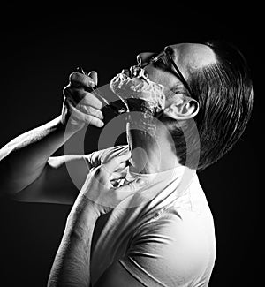 Black and white. Portrait of adult man in white t-shirt and glasses shaving with razor blades and shaving gel foam