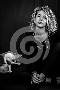 Black and white portrait of a beautiful young woman with blond curly hair sitting on the ground looking bored with a cat