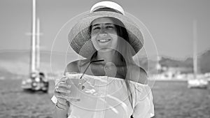 Black and white portrait of elegant woman standing with cocktail against calm sea and yachts at pier