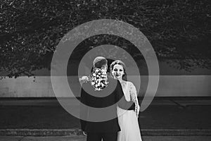 Black and white portrait of the groom, standing with his back, the bride hugging him holding flowers, looking at the camera