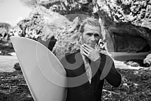 Black and white portrait of handsome man surfer in black wetsuit, holding white surf board  and blue ocean, cliff, rocks behind