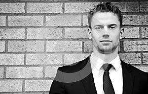 Black and white portrait of handsome man wearing suit standing next to brick wall looking at camera