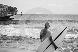 Black and white portrait of handsome shirtless man surfer , holding white surf board  and cliff, rocks on background