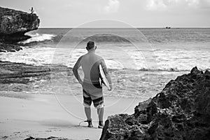 Black and white portrait of handsome shirtless man surfer , holding white surf board  and cliff, rocks, wave on background