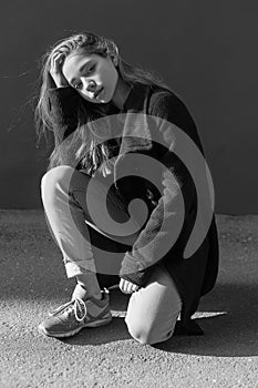 Black and white portrait of sad lonely young girl with long hair in coat, jeans