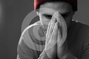 Black and white portrait of a young bearded man in a hat, hands clasped on his face, prays, his eyes down. Studio photo on a gray