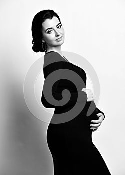 Black and white portrait of young beautiful happy pregnant woman in black elegant dress touching belly and looking at camera