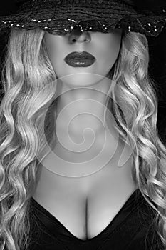 Black and white portrait of young blonde woman in black hat with black hat decollete and lush breasts, on a black background. photo