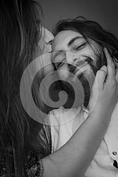 Black and white portrait of a young couple where a long haired woman kisses the man`s forehead while stroking his beard